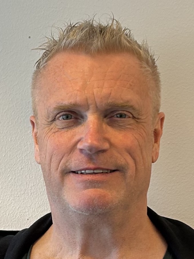 En medelålders man med kort, ljust hår är vänd mot kameran och ler lätt mot en vanlig, ljus bakgrund.
